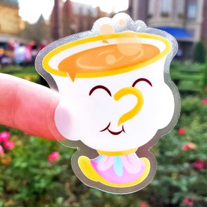 May include: A clear vinyl sticker of a cartoon teacup with a smiling face and a yellow handle. The teacup is filled with yellow liquid and has a pink and blue base.