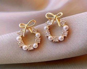 May include: A pair of gold-toned stud earrings. Each earring features a wreath design with faux pearls and clear crystals. A small bow sits atop each wreath. The earrings are displayed on a light gray fabric.