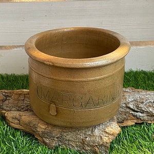 Może przedstawiać: Gliniany garnek w kolorze brązowym z napisem "MARGARINE" na boku, stojący na kawałku drewna unoszącego się na wodzie na tle zielonej sztucznej trawy.