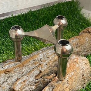 Peut inclure: Un bougeoir en argent à trois bras avec une base triangulaire, reposant sur deux morceaux de bois flotté. Le bougeoir est sur un lit d'herbe artificielle verte.
