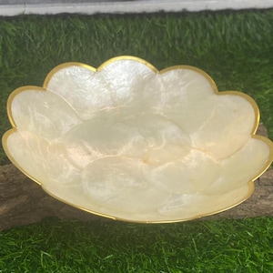Op de afbeelding: Een witte, decoratieve kom met een gouden rand. De kom is gemaakt van parelmoer en heeft een gegolfde rand. Hij staat op een stuk drijfhout.