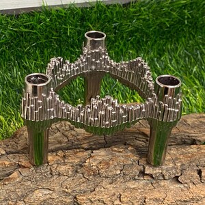 Può includere: Un portacandele in argento con tre bracci, ognuno con una piccola coppa per una candela. Il portacandele è realizzato in metallo testurizzato e ha un design moderno. È appoggiato su un pezzo di corteccia d'albero.