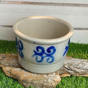 Puede incluir: Un pequeño macetero de cerámica blanca con diseños de espirales azules. El macetero está sentado sobre un trozo de madera a la deriva sobre un lecho de hierba artificial verde.