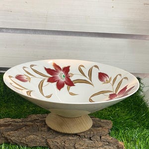 Op de afbeelding: Een witte keramische kom met een bloemenontwerp met rode en gouden bloemen. De kom staat op een houten standaard.