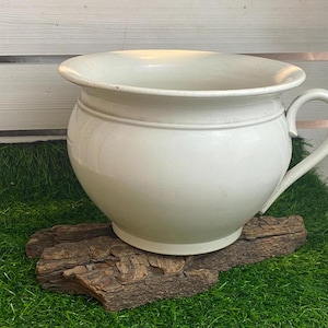 Op de afbeelding: Een witte keramische pot met een rond lichaam en een enkele handgreep. De pot staat op een stuk drijfhout.