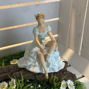 Peut inclure: Une figurine en porcelaine d'une ballerine portant une robe bleu clair et des chaussures blanches, assise sur une bûche entourée d'herbe verte et de marguerites blanches.