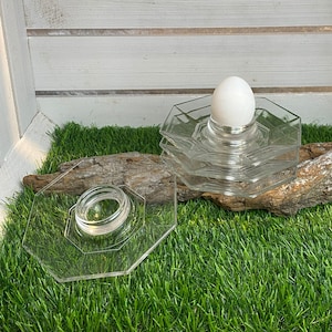Może przedstawiać: Białe jajko leży w przezroczystym szklanym stojaku na jajka. Stojak składa się z kilku warstw szkła i spoczywa na kawałku drewna unoszącego się na wodzie. Drewno unoszące się na wodzie leży na zielonej sztucznej trawie.