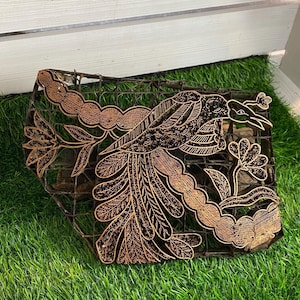 Könnte beinhalten: Ein brauner Holz-Batik-Stempel mit einem detaillierten Design eines Vogels und einem Blumenmuster. Der Stempel liegt auf einer grünen künstlichen Grasfläche.