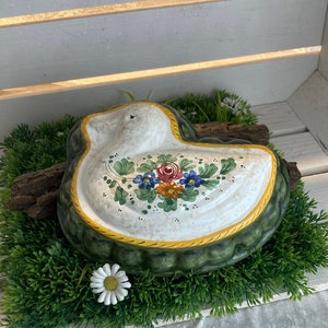 Könnte beinhalten: Eine weiße Keramikform in Form einer Ente mit grünen und gelben Akzenten und Blumendesigns. Die Form steht auf einem Bett aus grünem Kunstgras.
