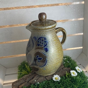 Op de afbeelding: Een bruine keramische kan met blauwe en witte bloemenmotieven staat op een bed van groen kunstgras met witte bloemen. De kan heeft een deksel en een handvat.