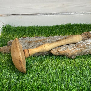 Może przedstawiać: Drewniany młotek z zaokrągloną główką i długim trzonkiem. Młotek jest jasnobrązowy i wydaje się być wykonany z jednego kawałka drewna. Spoczywa na kawałku kory i zielonej sztucznej trawie.