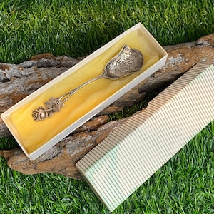 Peut inclure: Une cuillère à café en argent avec un motif de rose sur le manche, dans une boîte blanche avec une doublure jaune. La boîte est sur un morceau de bois flotté, qui est sur un lit d'herbe verte.