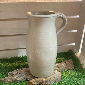 Peut inclure: Un pichet en céramique blanche avec une anse, posé sur un morceau de bois flotté sur un lit d'herbe artificielle verte.