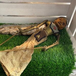 Könnte beinhalten: Eine Holzpuppe mit bemaltem Gesicht und brauner Stoffkleidung. Die Puppe liegt auf einem Bett aus grünem Kunstgras.