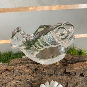 Może przedstawiać: Przezroczysta szklana figurka ptaka z precyzyjnymi detalami. Ptak siedzi na kawałku drewna, otoczony sztuczną trawą i białymi kwiatami stokrotki. Figurka ma teksturowaną powierzchnię i jest przedmiotem dekoracyjnym.