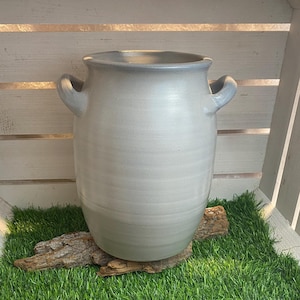 Könnte beinhalten: Eine weiße Keramikvase mit zwei Henkeln steht auf einem Stück Treibholz auf einem Bett aus Kunstgras. Die Vase befindet sich in einer weißen Holzkiste.