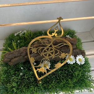 Może przedstawiać: Złoty, metalowy, sercowaty podkładka pod gorące z misternymi wzorami. Podkładka leży na sztucznej trawie w kolorze zielonym z białymi kwiatami.