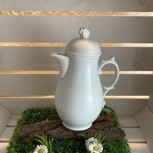 Può includere: Lattiera in porcellana bianca con coperchio e manico, appoggiata su un letto di erba artificiale verde con fiori di margherita bianchi.