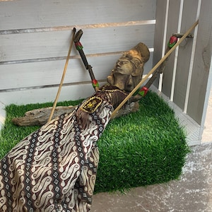 Könnte beinhalten: Eine Holzpuppe mit einem braunen und weißen gemusterten Stoffrock liegt in einer weißen Holzkiste mit grünem Gras darin. Die Puppe ist aus Holz und hat ein bemaltes Gesicht. Die Puppe hält einen Stock in jeder Hand.