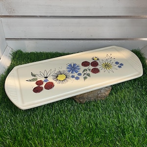 Peut inclure: Un plateau de service en céramique blanche avec un motif floral composé de fleurs rouges, jaunes, bleues et vertes. Le plateau est posé sur une surface de gazon artificiel vert.