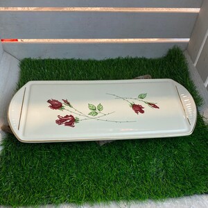 Puede incluir: Una bandeja de servir de cerámica blanca con un borde dorado y un diseño floral con rosas rojas. La bandeja está sobre una superficie de césped artificial verde.