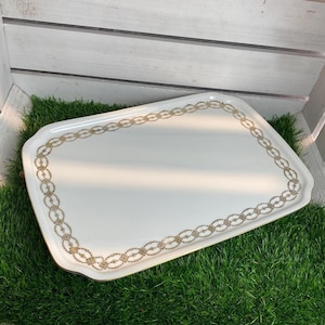 Puede incluir: Una bandeja rectangular blanca para servir con un borde decorativo dorado. La bandeja está sobre una superficie de césped artificial verde.