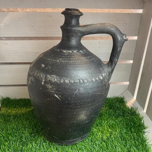 Könnte beinhalten: Eine schwarze Tonkrug mit Henkel und dekorativen Details. Der Krug steht auf einer grünen Kunstgrasoberfläche.