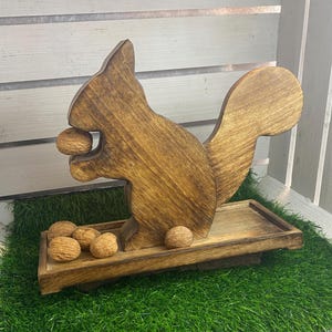Könnte beinhalten: Nussknacker in Form eines Eichhörnchens aus Holz mit einer Tablettbasis. Das Eichhörnchen hält eine Walnuss im Maul, mit weiteren Walnüssen im Tablett. Das Holz hat einen warmen, braunen Farbton.