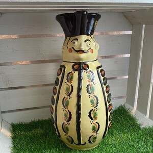 Può includere: Un'anfora in ceramica a forma di persona con un cappello nero, corpo giallo e dettagli dipinti in verde e arancio. L'anfora ha un viso sorridente con un baffo nero.