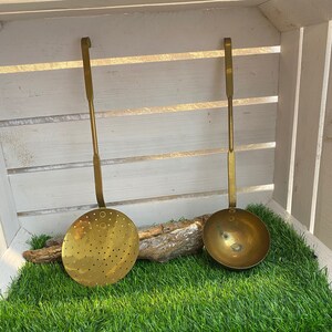 Può includere: Due utensili da cucina in ottone antico con manici lunghi. L'utensile a sinistra è un mestolo forato con una ciotola rotonda. L'utensile a destra è un mestolo con una ciotola rotonda. Entrambi gli utensili sono appesi a una cassa di legno.