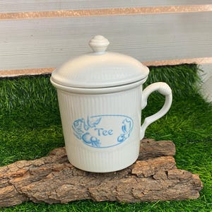 Könnte beinhalten: Weiße Teetasse aus Keramik mit blauem Blumendesign und dem Wort "Tee" in einem blauen Oval. Die Tasse hat einen Deckel.