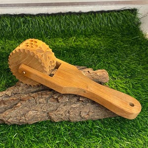 May include: A wooden massage roller with a floral pattern on the rolling surface. The handle is smooth and ergonomic. The roller is made of light-coloured wood and rests on a piece of bark, set against a backdrop of green grass.
