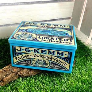 May include: A rectangular light blue metal tin featuring vintage-style graphics and text. The lid and front display the brand name J.G. KEMM, along with the text Kuchen-u. Zwieback-Fabrik, Lokstedt bei Hamburg, and the founding year 1702.