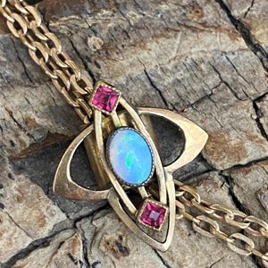 Peut inclure: Collier pendentif doré avec une opale ovale aux reflets bleus et verts, encadrée de deux pierres rouges carrées. Le pendentif de style Art nouveau est orné d'une chaîne.