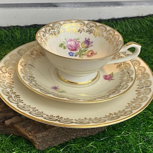 Può includere: Un servizio da tè vintage con tazza, piattino e piatto. La porcellana color avorio presenta finiture dorate e un motivo floreale con fiori rosa, viola e gialli. Il set è esposto su un pezzo di legno.