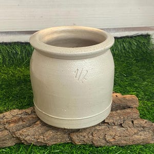 Puede incluir: Un tarro de gres gris claro con un cuerpo redondeado y una abertura ancha. El número "1/2" está estampado en la parte delantera. El tarro se encuentra sobre un trozo de corteza, con un fondo de hierba verde.