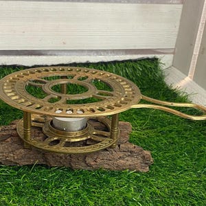 Könnte beinhalten: Ein messingfarbener Kerzenwärmer mit kreisförmigem Design und Griff. Der Wärmer steht auf einem Holzstück und hält eine kleine Kerze. Das Design umfasst dekorative Aussparungen und einen runden Sockel.