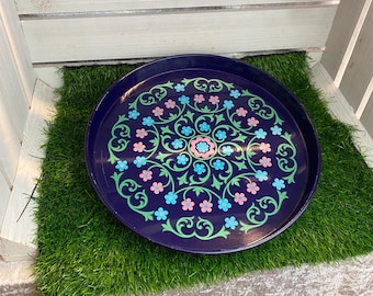 May include: A round, navy blue serving tray with a floral design. The pattern features pink, blue, and green flowers and vines arranged in a symmetrical pattern. The tray is placed on a patch of green artificial grass.