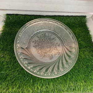 Könnte beinhalten: Transparenter Glasteller mit geprägtem Blumen- und Rankenmuster. Der Teller hat einen dekorativen Rand und ein zentrales Blumenmotiv. Der Text "Salz und Brot macht Wangen roth" ist am Rand geprägt.