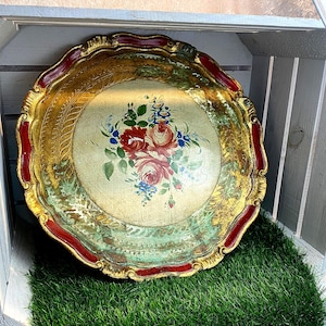 Könnte beinhalten: Ein rundes Dekotablett mit einem zentralen Blumenstrauß aus roten Rosen und blauen Blüten auf cremefarbenem Grund. Das Tablett hat einen gewellten Rand mit dunkelroter Umrandung und ein abgenutztes Blattgold-Finish mit grünen und braunen Akzenten.