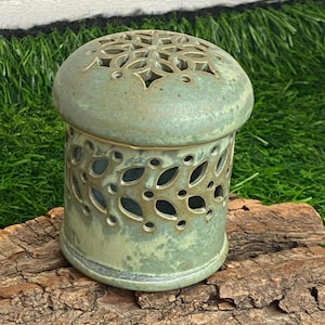 Peut inclure: Un photophore en céramique émaillée verte. Le corps cylindrique présente des découpes en forme de feuilles, et le couvercle en forme de dôme a un motif floral découpé. Le photophore est posé sur un morceau de bois.