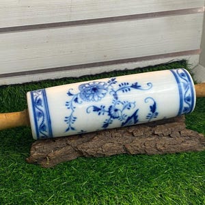 Può includere: Un mattarello con un corpo in ceramica bianca con motivi floreali blu e manici in legno. Il mattarello poggia su un pezzo di corteccia, su uno sfondo di erba artificiale verde e legno bianco.