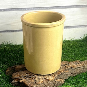 Pode incluir: Um pote de cerâmica cilíndrico com um acabamento liso, brilhante e de cor castanho-claro. O recipiente apresenta um design simples de paredes retas com uma borda ligeiramente alargada no topo, oferecendo uma aparência limpa e funcional.