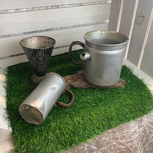 Könnte beinhalten: Drei antike Metallbehälter auf einer grünen Oberfläche. Dazu gehören ein hoher, kegelförmiger Becher mit Text, ein zylindrischer Becher mit Henkel und ein größerer Krug mit Ausguss und Henkel. Sie sind auf Kunstrasen angeordnet.