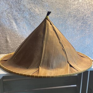 Può includere: Un cappello conico intrecciato di colore marrone con una cima appuntita e un fiocco nero. Il cappello ha un'ampia tesa ed è fatto di materiali naturali.