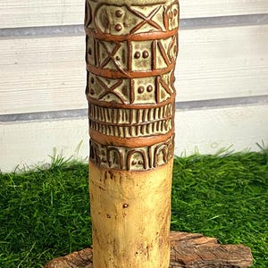 Könnte beinhalten: Eine hohe, zylindrische Keramikvase mit hellbrauner, matter Oberfläche. Der obere Teil ist mit geprägten geometrischen Mustern versehen, darunter Quadrate mit Kreuzen und Punkten, vertikale Linien und Bögen, die in dunkelrotbrauner Glasur abgesetzt sind.