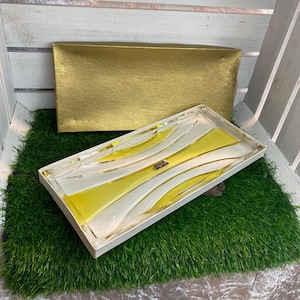 Peut inclure: Un plateau de service rectangulaire avec un motif de vagues blanches et jaunes, présenté dans une boîte blanche. Une boîte rectangulaire dorée est derrière le plateau. Le plateau est sur un lit d'herbe verte.