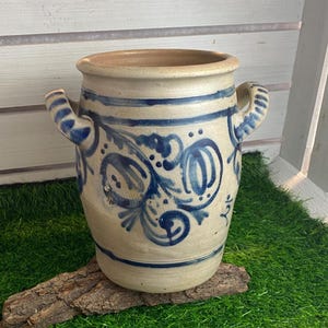 May include: A beige ceramic vase with blue floral patterns and two handles. The vase has a wide opening and is decorated with horizontal blue stripes. It sits on a piece of wood.