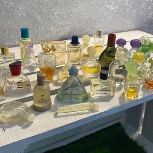 Puede incluir: Una colección de varios frascos de perfume de diferentes formas y tamaños. Los frascos son de vidrio transparente y presentan tapas y etiquetas de colores. Algunos frascos están vacíos, mientras que otros contienen líquido. Los frascos están dispuestos sobre una superficie blanca.