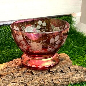 Könnte beinhalten: Eine tiefrote Glasschale mit einem geätzten Blumenmuster aus Blättern und Blüten, das die Außenseite umschließt. Die Schale hat einen breiten, abgerundeten Körper, der sich zu einem runden Boden hin verjüngt, mit glattem Rand und Oberfläche.